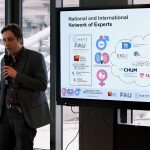 Man holding mike in hand and presenting ,while there is a big screen behind showing presentation