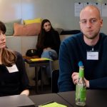 one man and woman are sitting and paying attention ,there i a table with laptop and bottle in front of them